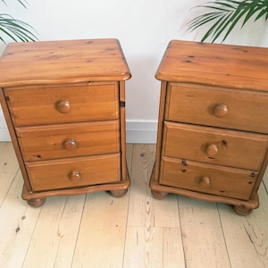May include: A pair of matching bedside cabinets crafted from light brown wood with a natural grain finish. Each cabinet features three stacked drawers, each equipped with a single round wooden knob, and rests on four small, rounded bun feet.