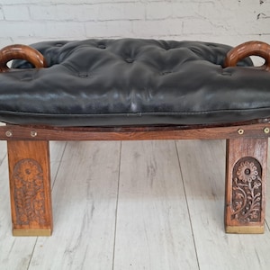 May include: A vintage footstool with a black leather cushion and wooden frame. The cushion is button-tufted. The wooden frame has carved floral designs on the legs and curved wooden armrests. A unique piece of furniture, approximately 100 cm long.