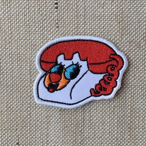May include: An embroidered patch depicting a rotary phone in white and red. The phone has a red receiver and a white base with black details. The phone has two large blue eyes and a small orange nose.