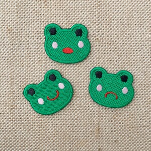 May include: Three green felt frog patches with embroidered faces. One frog has a red mouth and white eyes, one has a red smile and white eyes, and one has a blue frown and white eyes.