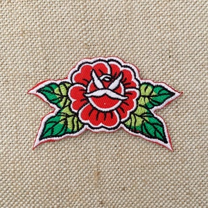 May include: Red embroidered rose with green leaves on a beige fabric background. The rose has a white center with a stylized bird design.