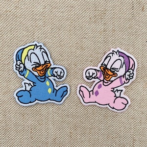 Op de afbeelding: Twee geborduurde patches met een cartoon eendje in babykleding. Eén is blauw met gele details, de andere roze met paarse stippen. Beide hebben oranje snavels en lachen.