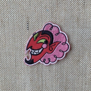 Pode incluir: Patch bordado com um diabo de desenho animado com rosto vermelho, cabelo preto e contorno rosa em forma de nuvem. O diabo tem um sorriso largo e olhos verdes. Para roupas ou acessórios.