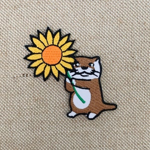 Könnte beinhalten: Ein braunes und weißes Otter, das eine gelbe Sonnenblume mit einem grünen Stiel hält. Das Otter ist auf einem beigefarbenen Stoff gestickt.