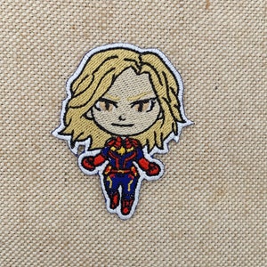 May include: A cartoon embroidered patch of Captain Marvel, a superhero with blonde hair, wearing a red, blue, and gold costume.