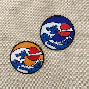 Pode incluir: Dois patches redondos bordados. Cada um apresenta um desenho de onda com um sol vermelho e um contorno branco. Um tem um fundo azul, o outro um fundo laranja. Ambos têm uma borda preta.