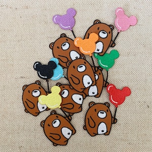 Pode incluir: Uma coleção de aplicações de ursos marrons segurando balões coloridos em forma de orelhas do Mickey Mouse. Os balões são de várias cores, incluindo vermelho, laranja, amarelo, verde, azul, roxo e preto.