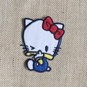 Op de afbeelding: Geborduurde strijkpatch met een cartoonfiguur met een wit gezicht, zwarte ogen, een rode strik en een blauwe overall. Het personage knippert met zijn oog en heeft een gele neus.