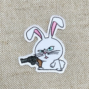 May include: White embroidered patch of a rabbit holding a gun with a serious expression. The rabbit has pink ears and blue eyes.