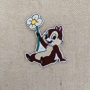May include: Embroidered patch featuring a cartoon chipmunk holding a white flower with a green stem. The chipmunk is brown with a pink bow on its head and is smiling.