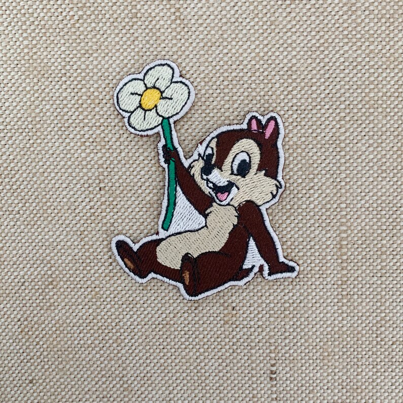 May include: Embroidered patch featuring a cartoon chipmunk holding a white flower with a green stem. The chipmunk is brown with a pink bow on its head and is smiling.