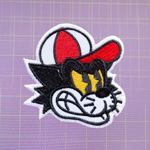 Op de afbeelding: Geborduurde strijkpatch van een cartoonkat met een rood-witte baseballpet. De kat heeft zwarte vacht, gele ogen en een wit gezicht. De kat fronste en heeft een chagrijnige uitdrukking.
