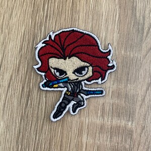 以下が含まれることがあります： 赤い髪、黒い目、白黒の衣装を着た漫画のキャラクターが刺繍されたパッチ。キャラクターは青と黄色の武器を持っています。