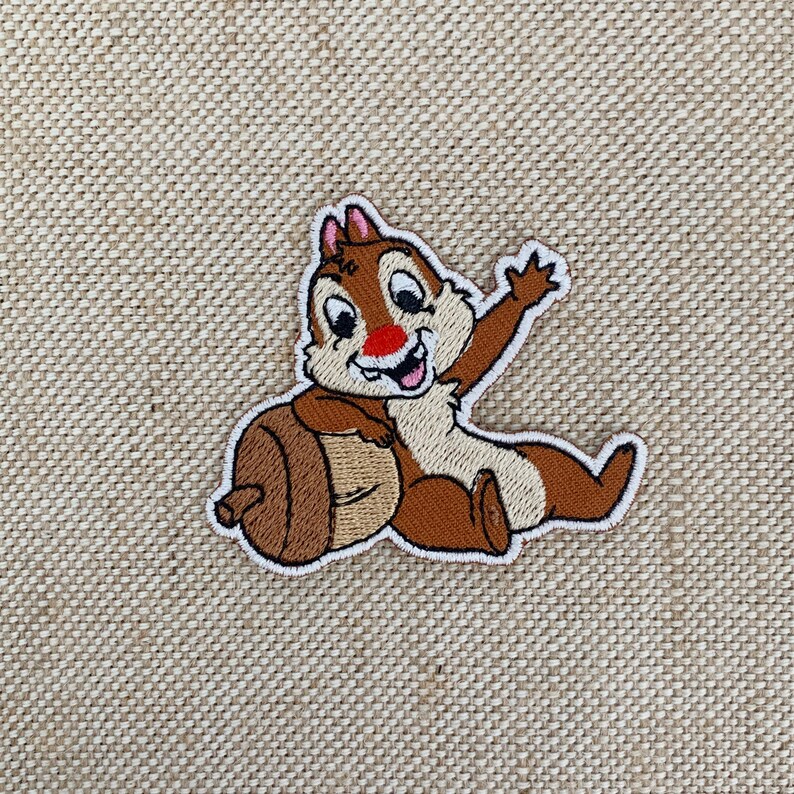 May include: An embroidered patch featuring Chip from Disney's Chip 'n Dale Rescue Rangers. Chip is sitting on an acorn and waving with his right paw.