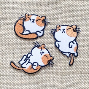 May include: Three embroidered iron-on patches featuring cartoon cats with orange and white fur. The cats are in different poses, one is sleeping, one is sitting up, and one is lying on its side.
