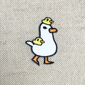 May include: White embroidered patch of a goose with two yellow ducklings on its back. The goose is walking with its legs outstretched and the ducklings are perched on its back and head.