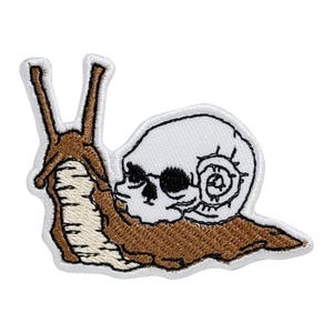 Op de afbeelding: Een geborduurde strijkpatch met een bruine slak met een witte schedel als kop. De slak kruipt naar rechts. De schedel heeft zwarte ogen en een gedetailleerde spiraalvormige schelp.