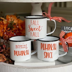May include: Three white enamel mugs with black rims and orange lettering. The mugs are stacked on a white tiered tray. The mugs say "Fall Sweet Fall", "Morning, Pumpkin", and "Pumpkin Spice".