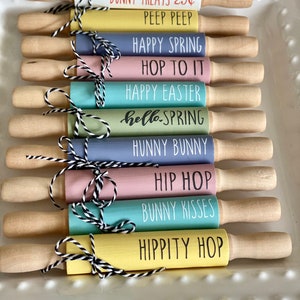 May include: Nine colorful rolling pins with different Easter-themed sayings. The sayings include "Bunny Treats 25c Peep Peep", "Happy Spring Hop To It", "Happy Easter", "hello-SPRING", "Hunny Bunny", "Hip Hop", "Bunny Kisses", and "Hippity Hop".