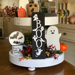 May include: A white tiered tray decorated with Halloween themed items. The tray features a black sign with the word "BOO" in white letters, a white ghost, a black bat, a brown pumpkin, and a white mug with the text "Black Cat Lady".