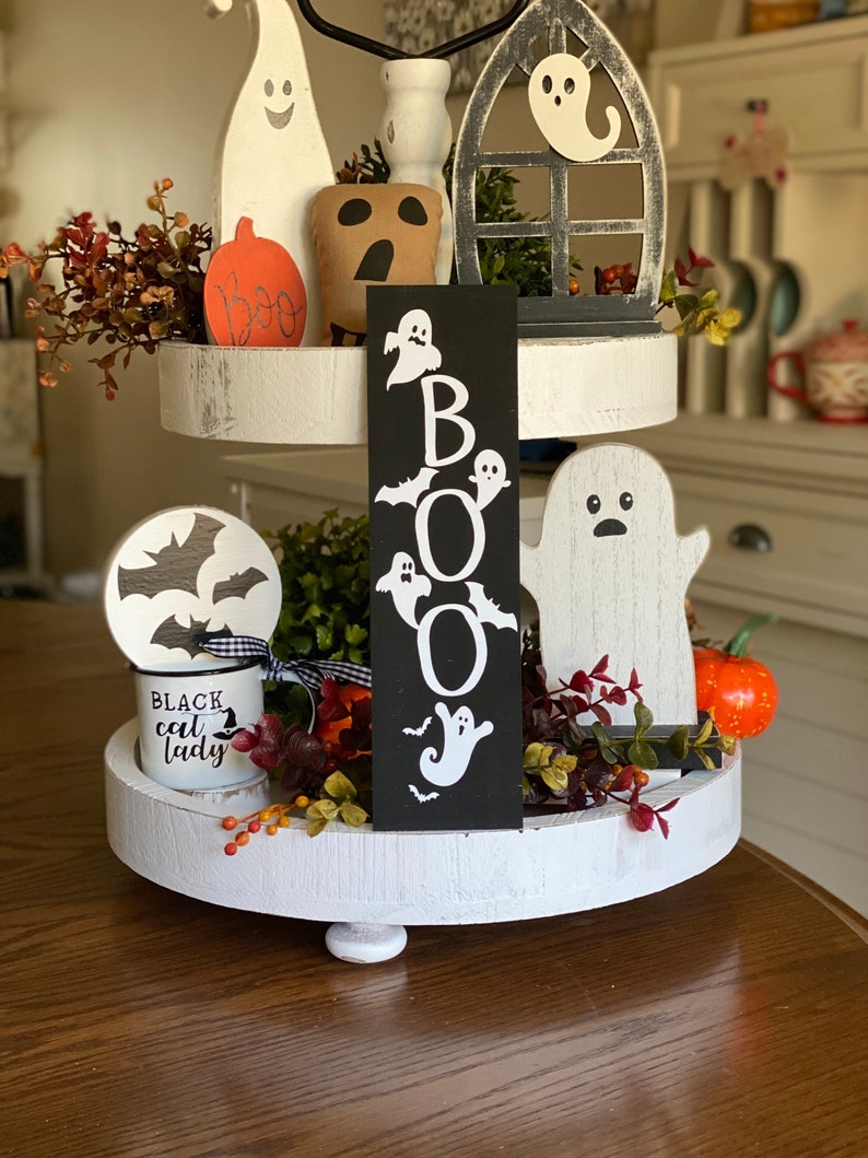 May include: A white tiered tray decorated with Halloween themed items. The tray features a black sign with the word "BOO" in white letters, a white ghost, a black bat, a brown pumpkin, and a white mug with the text "Black Cat Lady".