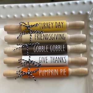May include: Five wooden rolling pins with different Thanksgiving-themed phrases painted on them. The phrases are "Turkey Day", "Friendsgiving", "Gobble Gobble", "Give Thanks", and "Pumpkin Pie".