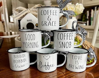May include: Six white enamel mugs with black rims and black lettering. The mugs have various coffee-themed sayings, including "Coffee & Grace", "Good Morning", "Coffee Snob", "Bibbidi Bobbidi Brew", "Love is Brewing", and "Coffee".