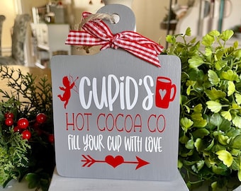 May include: A gray wooden sign with the text "Cupid's Hot Cocoa Co. Fill your cup with love" in white lettering. The sign has a red heart and a red arrow with a red heart in the center. The sign is decorated with a red and white gingham ribbon.