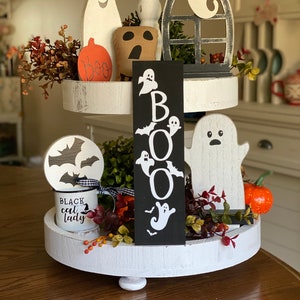 May include: A two-tiered white tiered tray decorated for Halloween with a black sign that says "BOO" with white ghosts and bats, a white ghost figurine, a black and white mug that says "Black cat lady", a white ghost with a black face, a brown ghost with a black face, and a red and orange pumpkin.