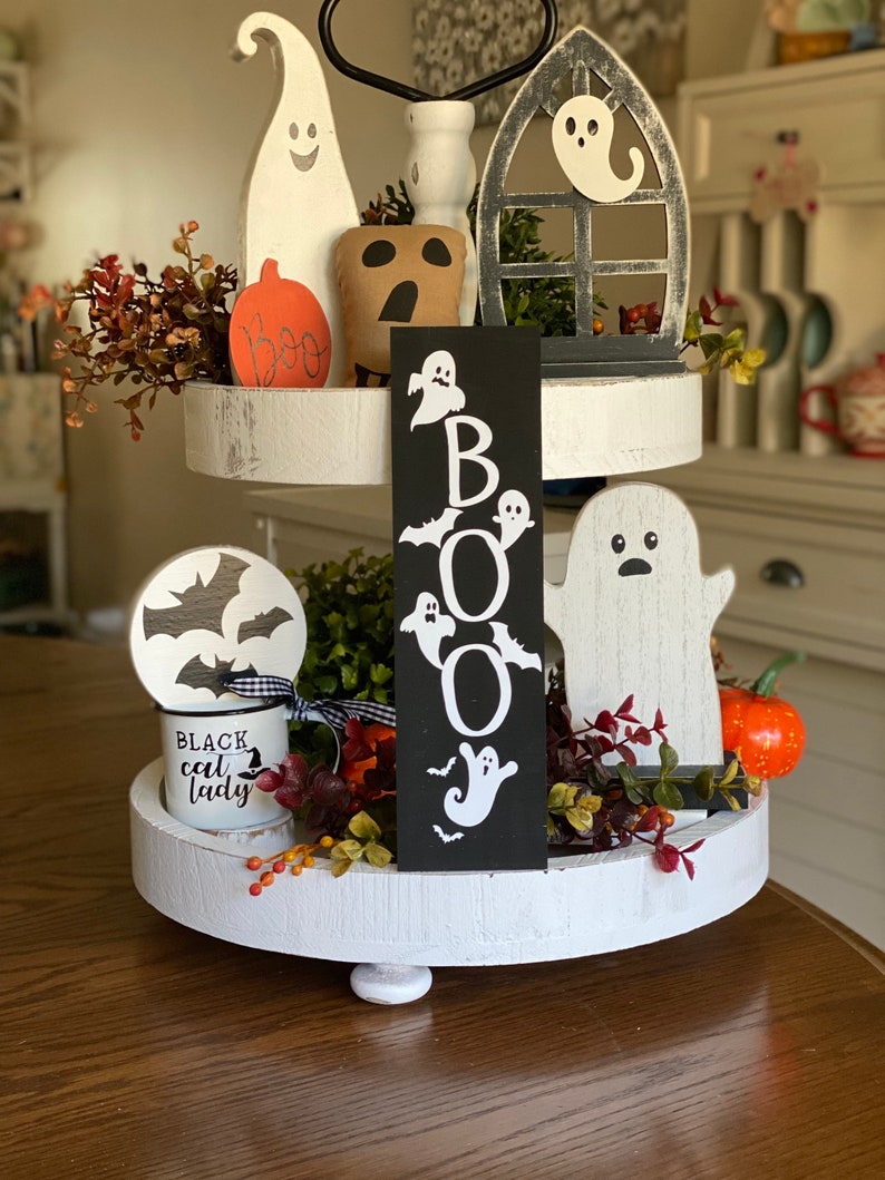 May include: A two-tiered white tiered tray decorated for Halloween with a black sign that says "BOO" with white ghosts and bats, a white ghost figurine, a black and white mug that says "Black cat lady", a white ghost with a black face, a brown ghost with a black face, and a red and orange pumpkin.