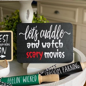 May include: Black chalkboard sign with white text that reads "let's cuddle and watch scary movies".  Two wooden rolling pins with green and black fabric and white text that reads "Feelin' Wicked" and "Broom Parking".