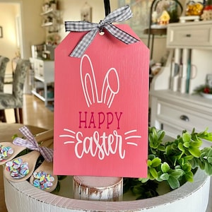 May include: Pink wooden sign with a white bunny ear design and the text "Happy Easter" in white lettering. The sign is hanging from a black hook and has a grey and white checked ribbon.