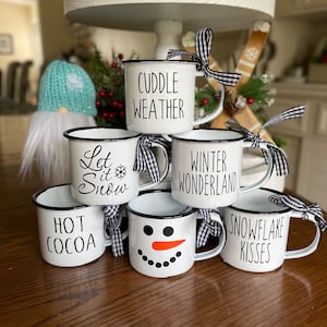 May include: Six white enamel mugs with black rims and black lettering. The mugs are stacked in a pyramid shape. The mugs have the following text: "Cuddle Weather", "Let it Snow", "Winter Wonderland", "Hot Cocoa", "Snowflake Kisses", and a snowman face.