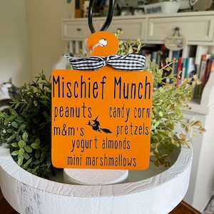 May include: Orange wooden sign with black text that reads "Mischeif Munch" and lists various Halloween treats including peanuts, candy corn, m&m's, pretzels, yogurt almonds, and mini marshmallows. The sign is hanging from a black metal hook and has a black and white checkered ribbon.