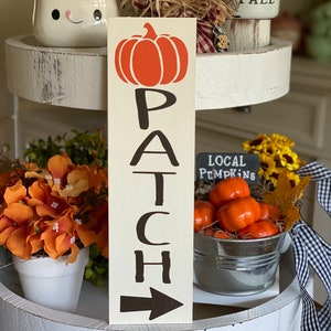 May include: A wooden sign with the word "PATCH" written vertically in black letters. An orange pumpkin is above the word. A black arrow points to the right at the bottom of the sign.