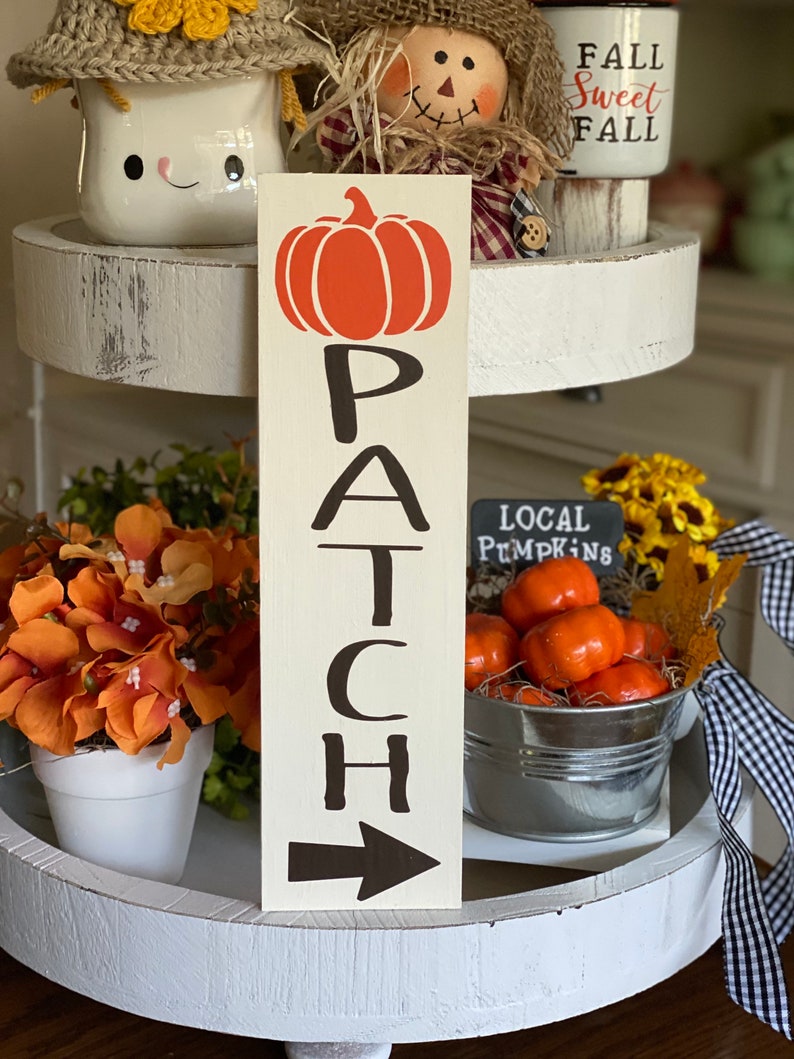 May include: A wooden sign with the word "PATCH" written vertically in black letters. An orange pumpkin is above the word. A black arrow points to the right at the bottom of the sign.