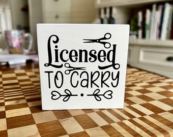 Puede incluir: Un letrero de madera blanco con texto negro que dice "Licensed to Carry" con un par de tijeras sobre la palabra "Licensed" y un par de tijeras debajo de la palabra "to".