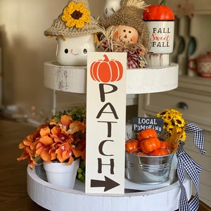 May include: A white tiered tray decorated with fall-themed items, including a pumpkin sign that reads "PATCH", a scarecrow, a mug with a crocheted sunflower hat, a pumpkin-shaped mug that says "Fall Sweet Fall", and a small metal bucket filled with orange pumpkins and a sign that says "LOCAL PUMPKINS".