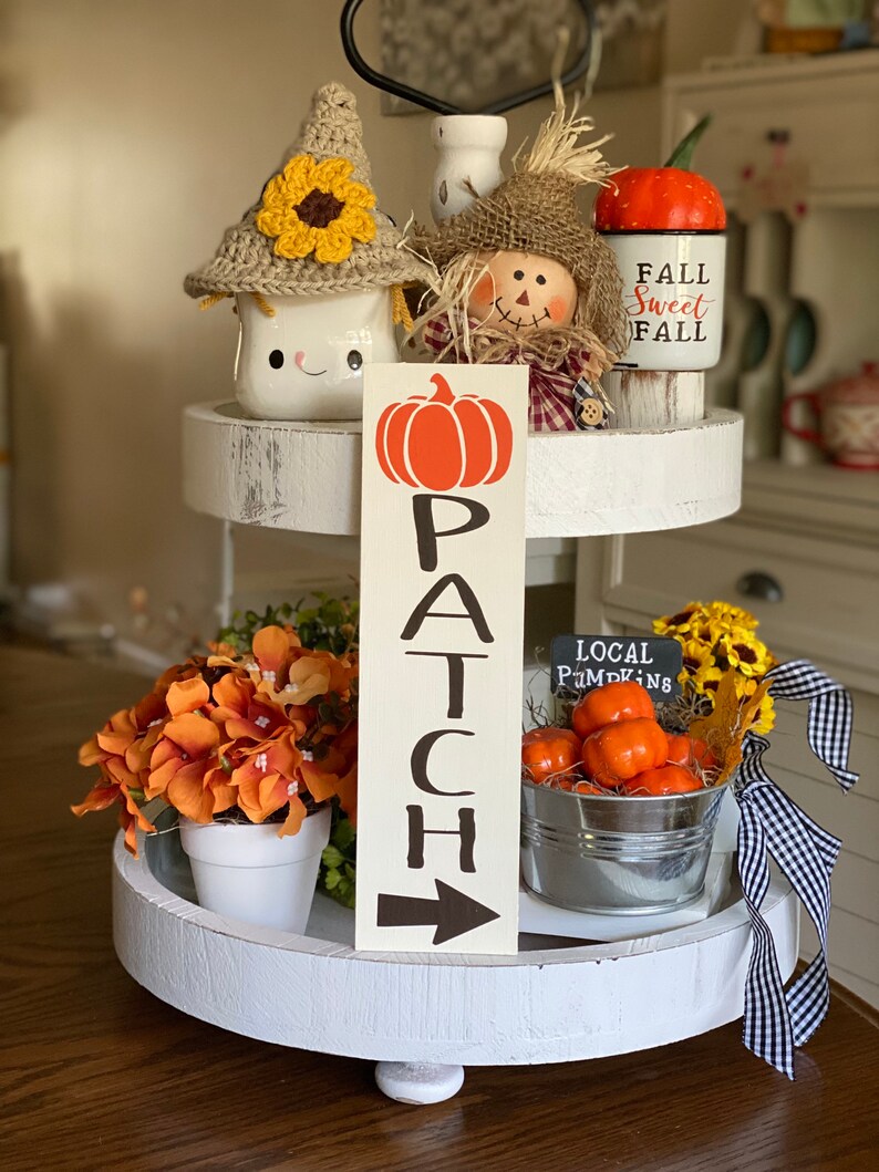 May include: A white tiered tray decorated with fall-themed items, including a pumpkin sign that reads "PATCH", a scarecrow, a mug with a crocheted sunflower hat, a pumpkin-shaped mug that says "Fall Sweet Fall", and a small metal bucket filled with orange pumpkins and a sign that says "LOCAL PUMPKINS".