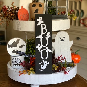 May include: A three-tiered white tiered tray decorated for Halloween. The top tier features a black and white ghost sign with the word "BOO" spelled out with ghosts and bats. The middle tier has a white ghost, a black and white bat, and a small orange pumpkin. The bottom tier has a black and white mug with the words "Black Cat Lady" and a small bouquet of fall foliage.