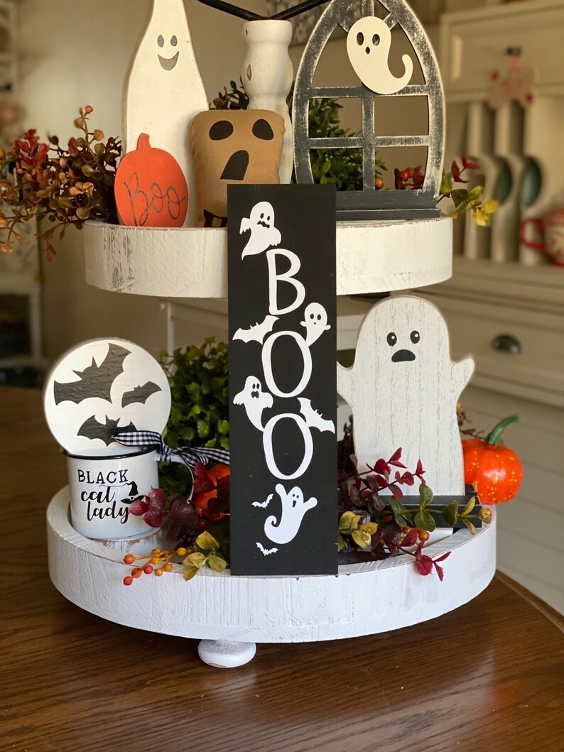 May include: A three-tiered white tiered tray decorated for Halloween. The top tier features a black and white ghost sign with the word "BOO" spelled out with ghosts and bats. The middle tier has a white ghost, a black and white bat, and a small orange pumpkin. The bottom tier has a black and white mug with the words "Black Cat Lady" and a small bouquet of fall foliage.
