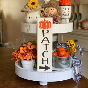 May include: A white tiered tray decorated with fall-themed items, including a pumpkin-shaped sign that reads "PATCH", a scarecrow figurine, a mug with a crocheted sunflower hat, and a small bucket filled with orange pumpkins. The sign also has an arrow pointing to the right.