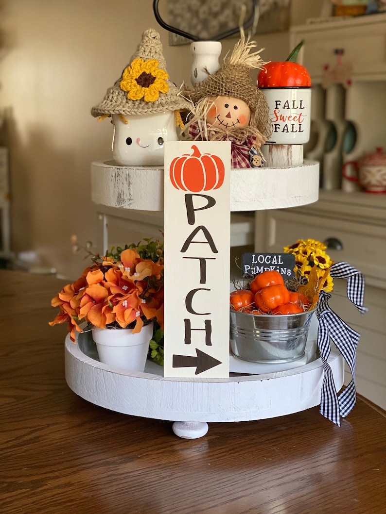 May include: A white tiered tray decorated with fall-themed items, including a pumpkin-shaped sign that reads "PATCH", a scarecrow figurine, a mug with a crocheted sunflower hat, and a small bucket filled with orange pumpkins. The sign also has an arrow pointing to the right.