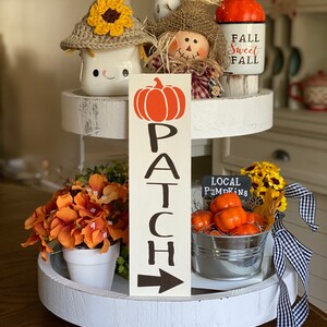 May include: A white tiered tray decorated with fall-themed items, including a crocheted snowman mug, a scarecrow figurine, a pumpkin-shaped mug with the text "Fall Sweet Fall", a wooden sign with the text "PATCH" and an arrow, a small pot of orange flowers, and a metal bucket filled with small orange pumpkins with a chalkboard sign that reads "LOCAL PUMPKINS".