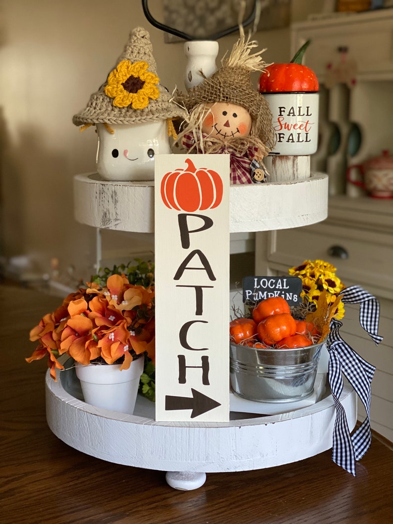 May include: A white tiered tray decorated with fall-themed items, including a crocheted snowman mug, a scarecrow figurine, a pumpkin-shaped mug with the text "Fall Sweet Fall", a wooden sign with the text "PATCH" and an arrow, a small pot of orange flowers, and a metal bucket filled with small orange pumpkins with a chalkboard sign that reads "LOCAL PUMPKINS".
