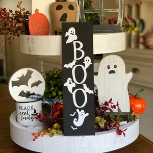 May include: A three-tiered white tiered tray decorated with Halloween themed items. The top tier features a black and white ghost, a black and white window, and a brown and white ghost. The middle tier features a black and white ghost, a black and white bat, a black and white mug with the text "Black Cat Lady", and a black sign with the word "BOO" in white letters. The bottom tier features a white ghost, a pumpkin, and greenery.