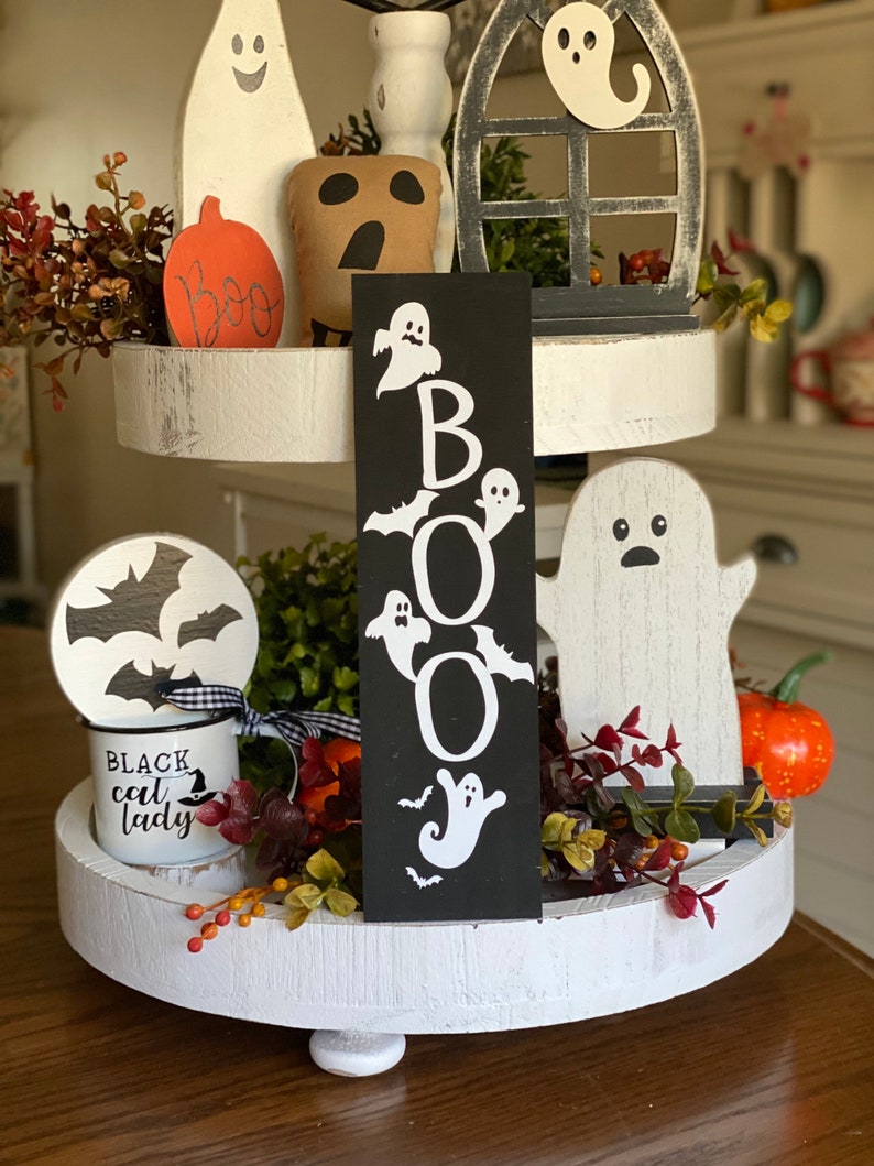 May include: A three-tiered white tiered tray decorated with Halloween themed items. The top tier features a black and white ghost, a black and white window, and a brown and white ghost. The middle tier features a black and white ghost, a black and white bat, a black and white mug with the text "Black Cat Lady", and a black sign with the word "BOO" in white letters. The bottom tier features a white ghost, a pumpkin, and greenery.