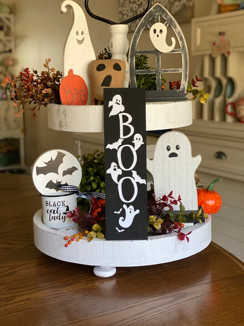 May include: A tiered tray decorated for Halloween with a black sign that says "BOO" with white ghost and bat cutouts, a white ghost figurine, a white ceramic mug with black text that says "Black cat lady", a white ghost figurine with a black face, a brown ghost figurine, a white ghost figurine in a black window frame, and a small orange pumpkin.