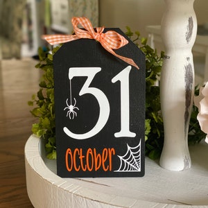 May include: Black wooden sign with white lettering that says "31 October" with a white spider and web design. The sign has a orange and white checkered ribbon tied around it.