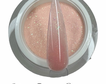 Könnte beinhalten: Nahaufnahme eines Glases mit nude-farbenem Glitzer-Nagellack. Das Glas ist weiß mit einem silbernen Deckel. Der Nagellack ist hellrosa mit schimmerndem Glitzer.
